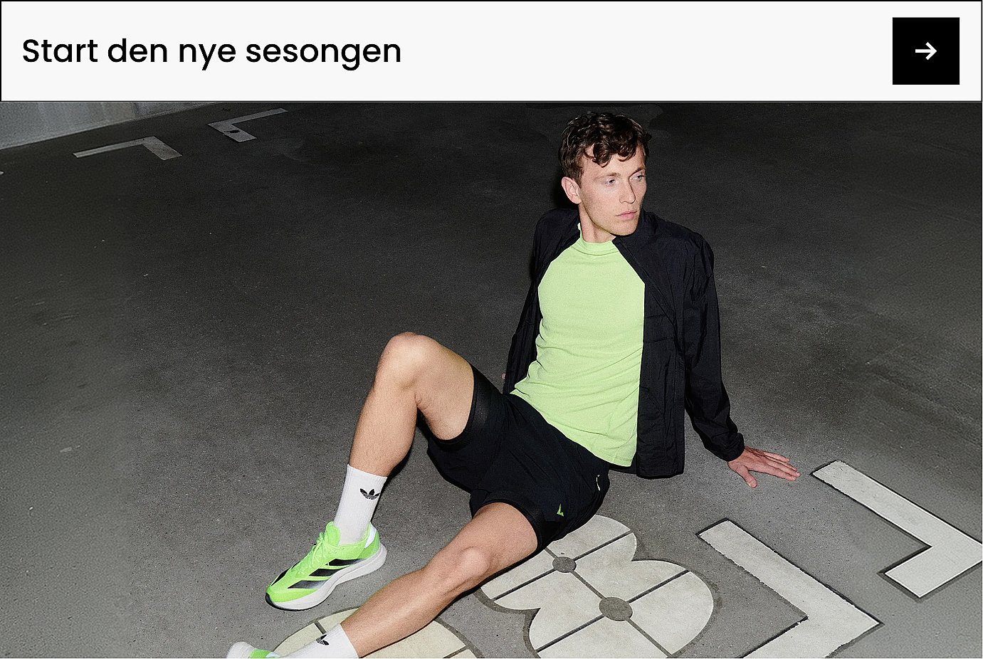 En mann i neongrønn treningstrøye og -sko, svart jakke og shorts, som sitter på en mørk betongflate med hvite markeringer. Sport.