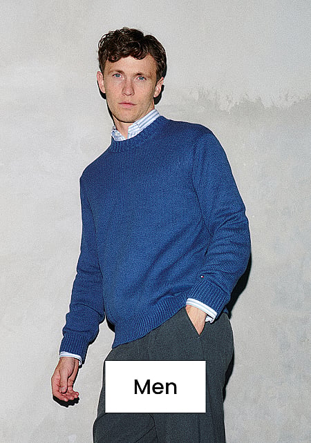 Man in blue sweater, striped shirt, and dark pants, standing against a gray wall. Text "Men" at bottom.