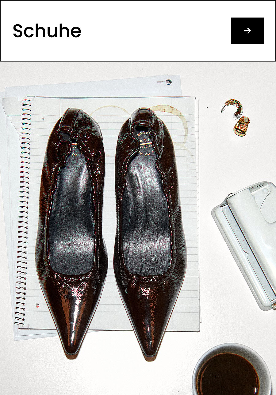 Ein Paar dunkelbraune Ballerinas aus Lackleder mit spitzer Zehenpartie auf einem Schreibtisch, auf dem ein Spiralblock, Kaffee, ein Locher und Ohrringe liegen. Text: Schuhe