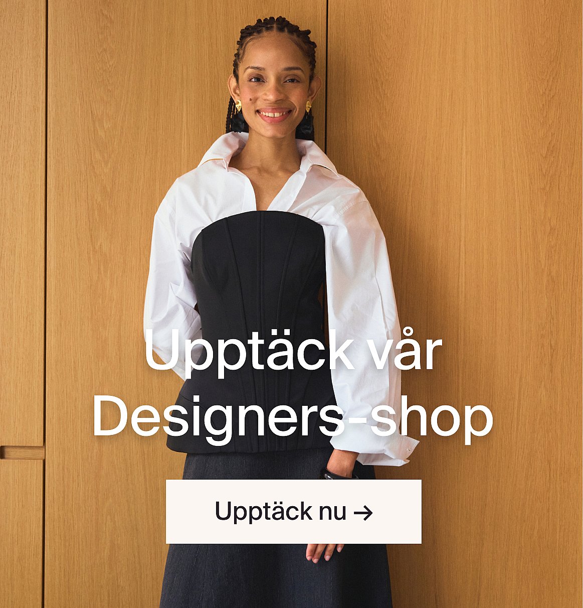 En leende kvinna i svart korsett och vit skjorta framför en trävägg. Text: Upptäck vår designbutik.