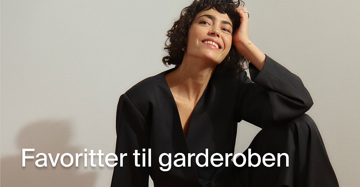 En smilende kvinde i en sort blazer, med hånden i sit krøllede hår. Tekst: Garderobeklassikere.