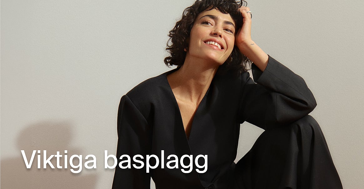 En leende kvinna i en svart kavaj, med handen i sitt lockiga hår. Text: Grundplagg.