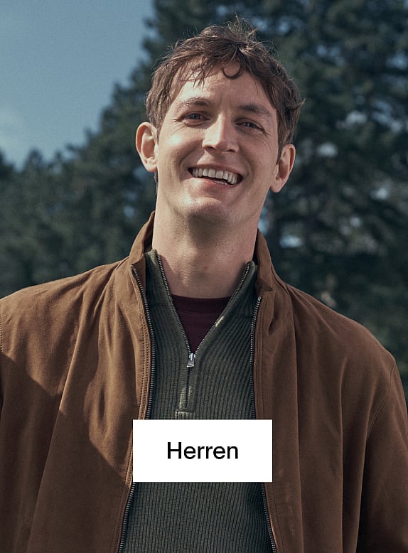 Ein lächelnder Mann in brauner Jacke und grünem Pullover im Freien, im Hintergrund Bäume. Der Text „Men“ auf einem weißen Kasten.