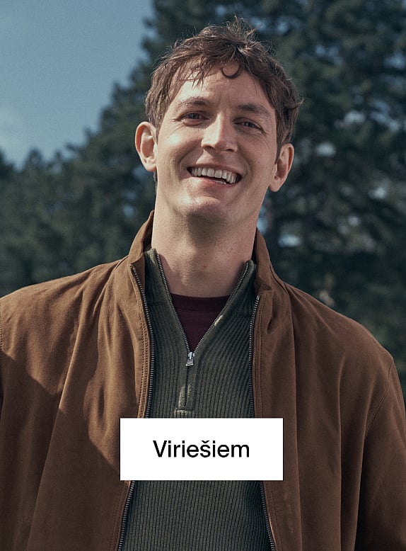 Smaidošs vīrietis brūnā jakā un zaļā džemperī ārā, fonā redzami koki. Uz balta laukuma uzraksts „Vīrieši”.
