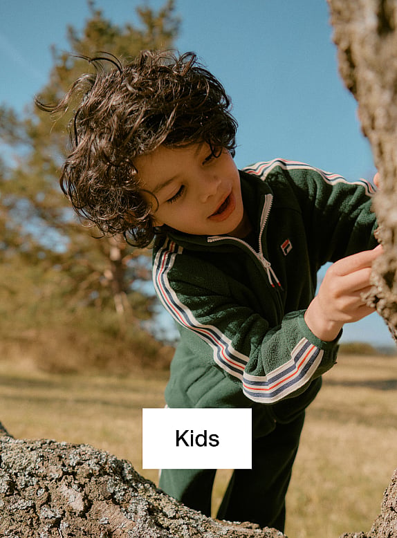 Curly-haired child in a green striped jacket, looking down and touching a tree trunk outdoors.