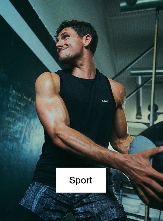 Ein muskulöser Mann in einem schwarzen Tanktop und gemusterten Shorts, der in einem Fitnessstudio einen Medizinball hält.