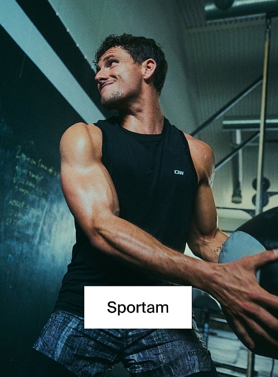 Muskuļots vīrietis melnā krekliņā bez piedurknēm un rakstainos šortos, sporta zālē turot medicīnas bumbu.