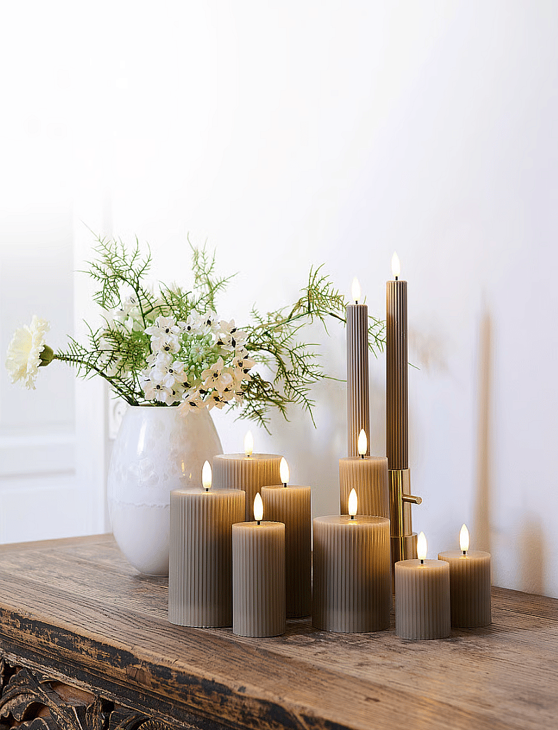 Lit grey ribbed candles and a white vase with flowers on a rustic wooden table against a light wall.