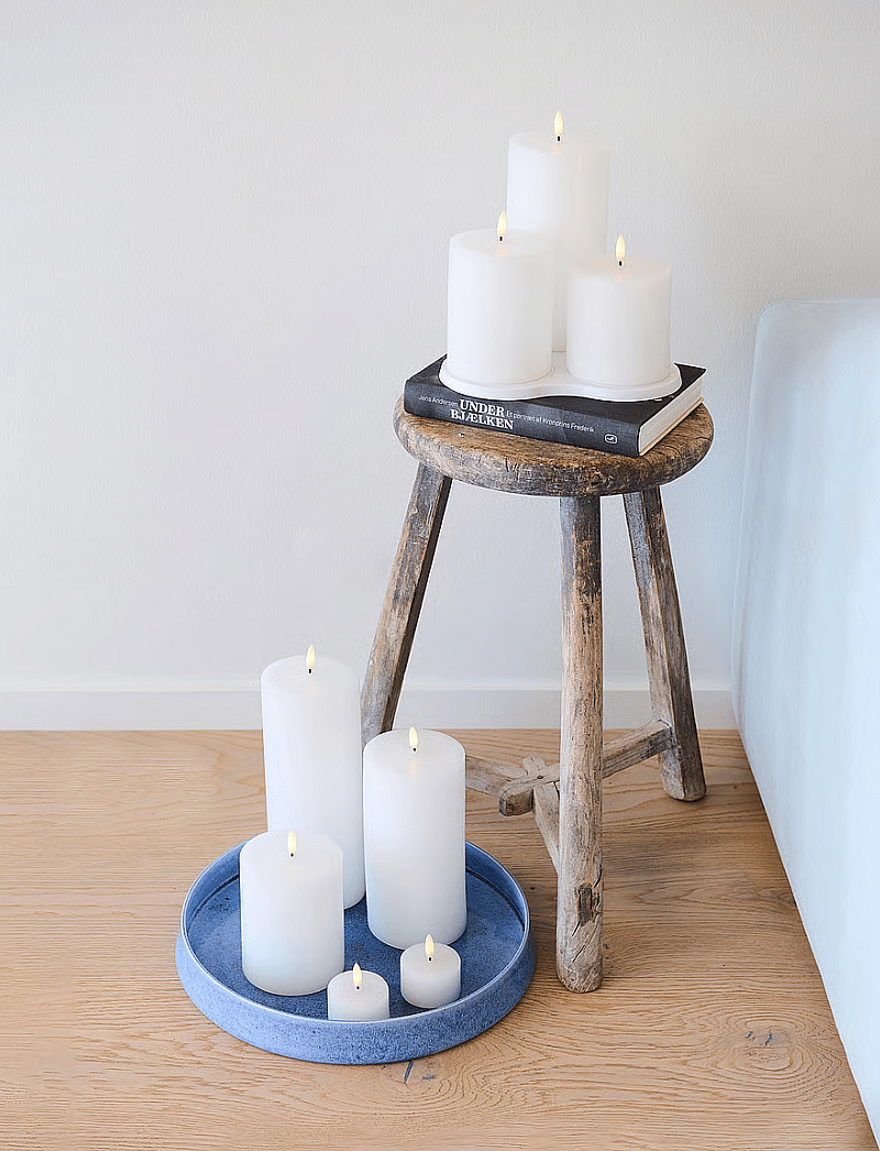 White candles on a rustic wooden stool with a book, and on a blue tray on a light wood floor.