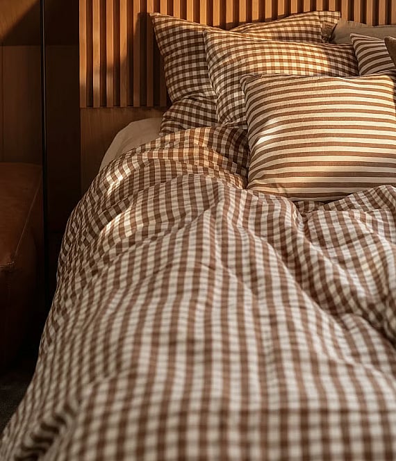 Ein Bett mit einer braun-weiß karierten Bettdecke und Kissen – eines davon gestreift – vor einem Kopfteil aus Holzlatten.