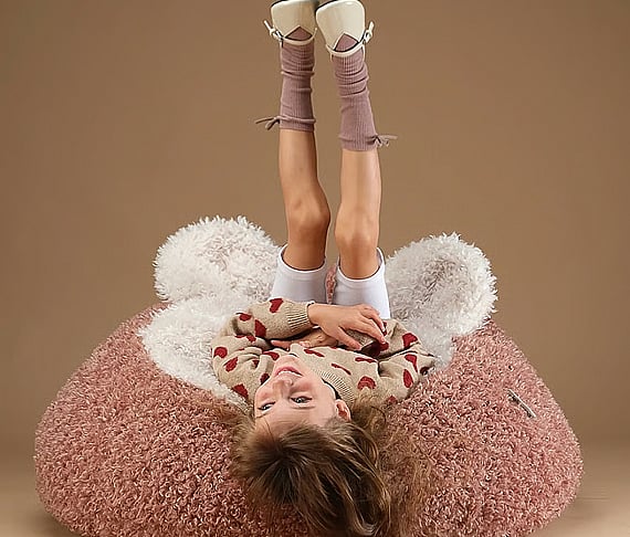 Smiling child upside down on a fluffy pink and white bear-shaped beanbag, wearing a heart sweater, white shorts, and pink