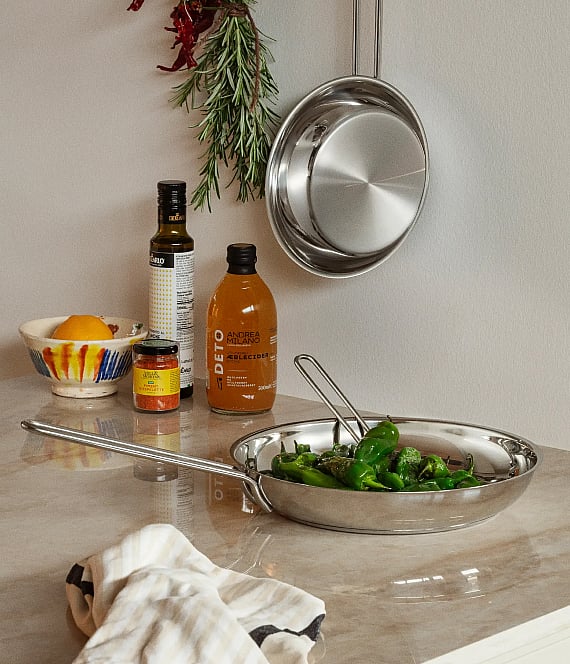 Eine Edelstahlpfanne mit grünen Paprikaschoten auf einer Küchenarbeitsplatte. Olivenöl, Apfelwein, Zitronen sowie eine hängende Pfanne und Rosmarin sind