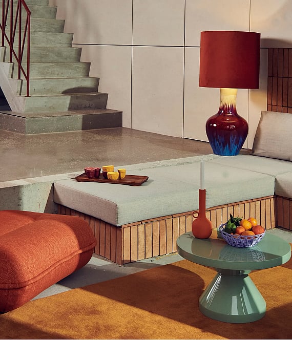 Modernes Wohnzimmer mit einer großen roten Lampe, einem grünen Sofa, einer orangefarbenen Ottomane, einem grünen Couchtisch und einer Betontreppe.