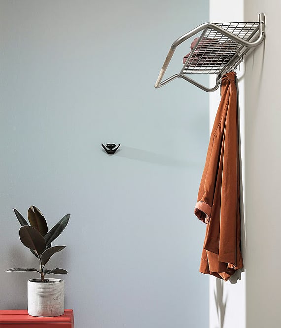 Eine hellblaue Wand mit einer Garderobe aus Metall, an der eine braune Jacke hängt, ein schwarzer Haken und eine Topfpflanze auf einem roten Sockel.