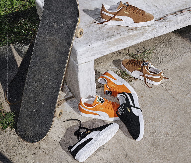 Skateboard leaning on a concrete bench, surrounded by six pairs of Puma suede sneakers in brown, orange, and black on
