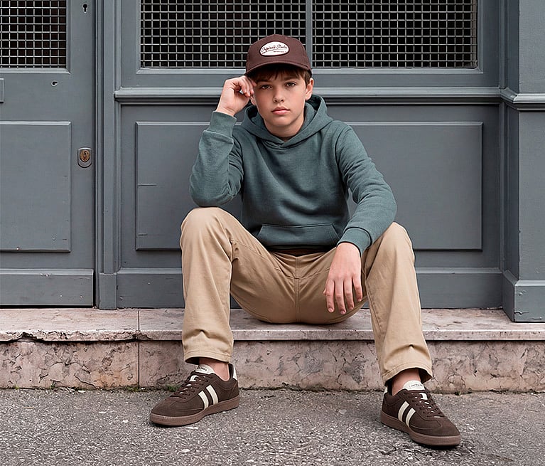Boy in brown cap, green hoodie, khaki pants, and brown sneakers sits on a stone step by a gray door.