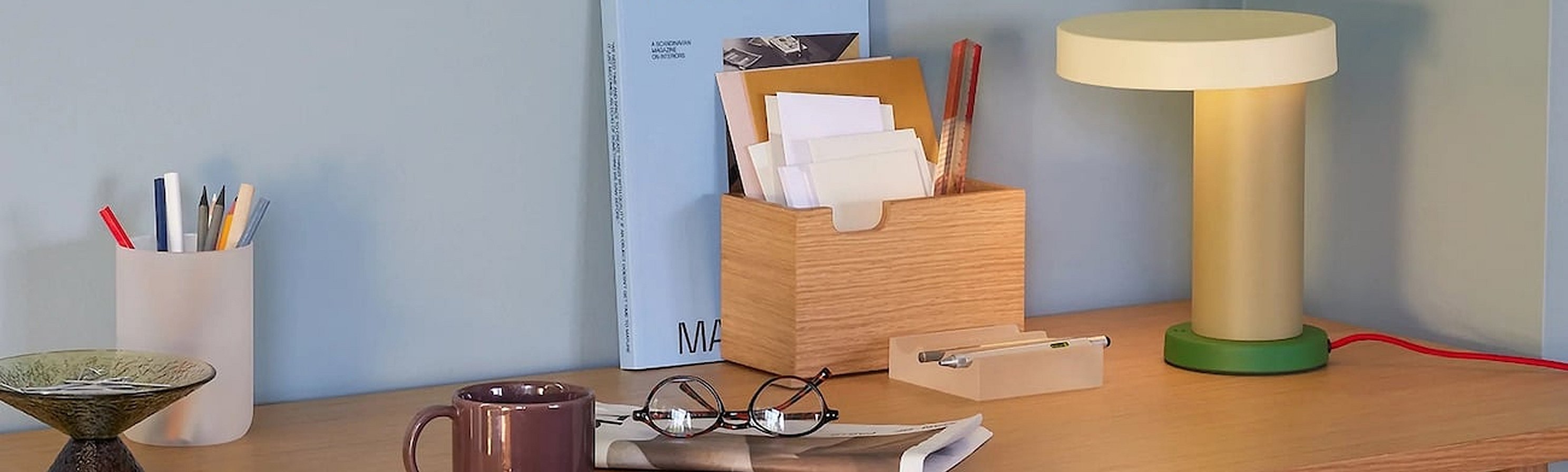 A light wood desk holds a cream and green lamp, wooden organizer, book, pen holder, mug, glasses, and dish against a blue