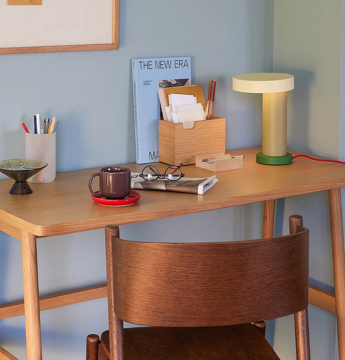Ein Schreibtisch im Homeoffice mit einer modernen Lampe, einer Kaffeetasse, Gläsern und Schreibutensilien. Im Vordergrund steht ein Holzstuhl.