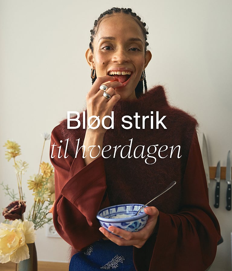 En smilende kvinde i en rød strikbluse, der spiser et bær, mens hun holder en skål i et køkken. Tekst: Bløde strikvarer til hverdagen.
