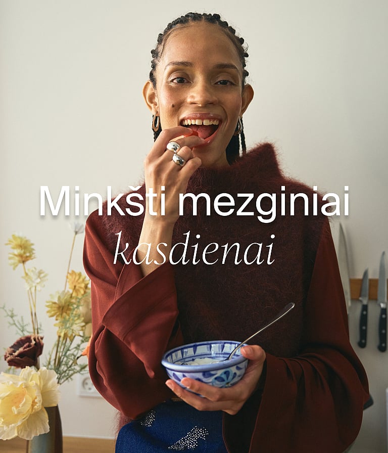 Šypsosi moteris, vilkinti raudoną megztinį, valgo uogą ir laiko dubenėlį virtuvėje. Tekstas: Minkšti megztiniai kasdienai.