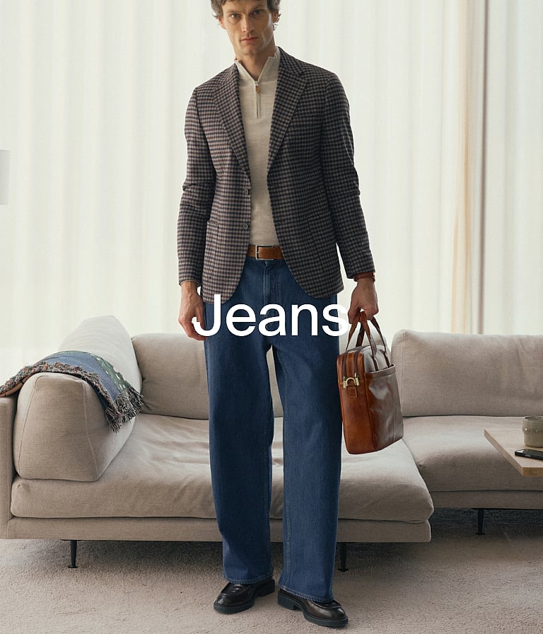 En man i rutig kavaj, ljus tröja, blå jeans och svarta skor, som håller i en brun portfölj och står i ett vardagsrum