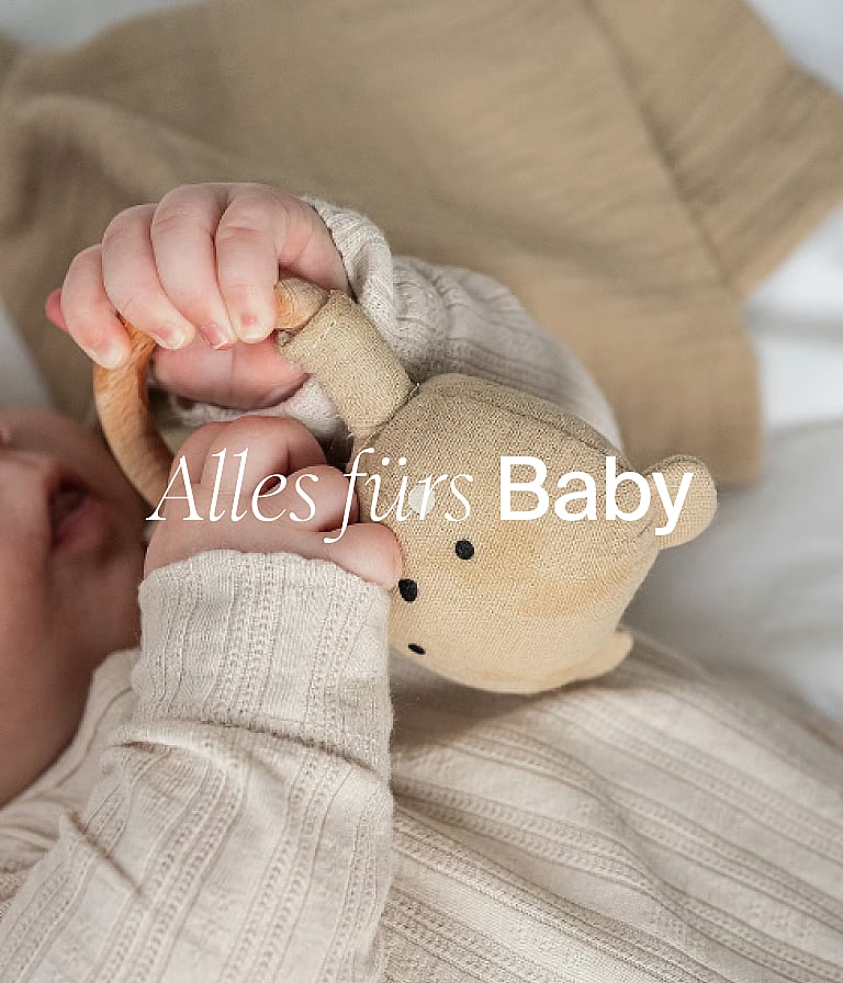 Die Hände eines Babys in schlichter Kleidung, die ein Stoffbären-Spielzeug halten. Bildunterschrift: „Baby-Grundausstattung“