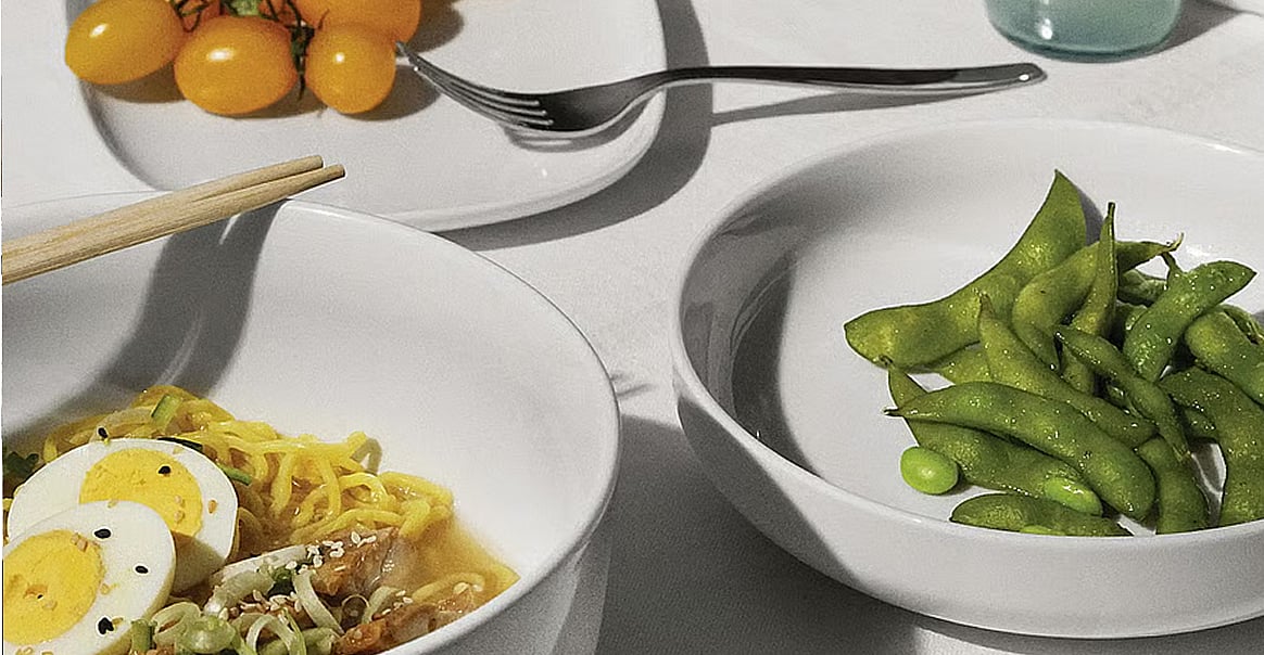 A table with a bowl of ramen and chopsticks, a bowl of edamame, and a plate of yellow cherry tomatoes with a fork.