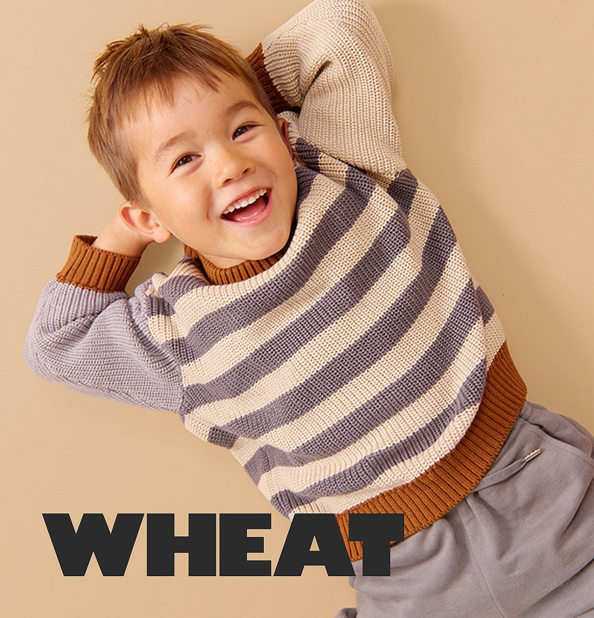 Smiling young boy lying on a beige background, wearing a striped sweater with hands behind his head.