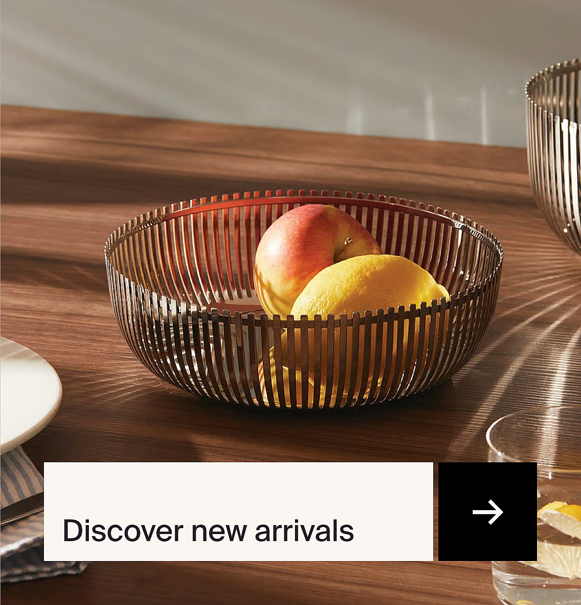 A slatted metal fruit bowl with an apple and lemons on a wooden table, with sunlight creating striped shadows.