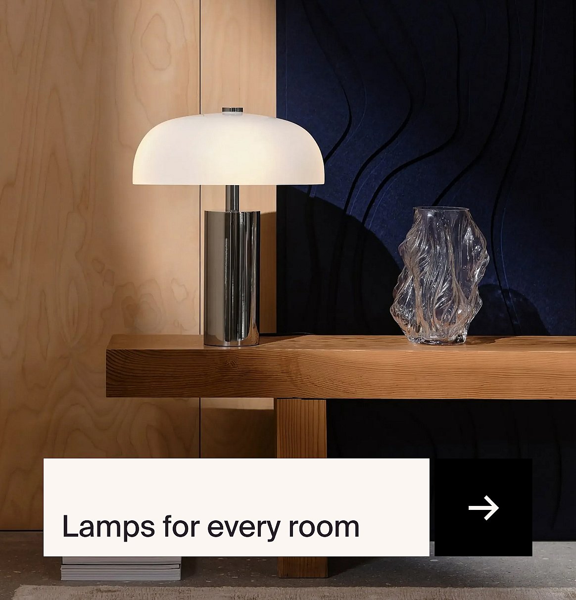 Modern table lamp with white mushroom shade and chrome base on a wooden shelf, next to a clear vase, against wood and dark