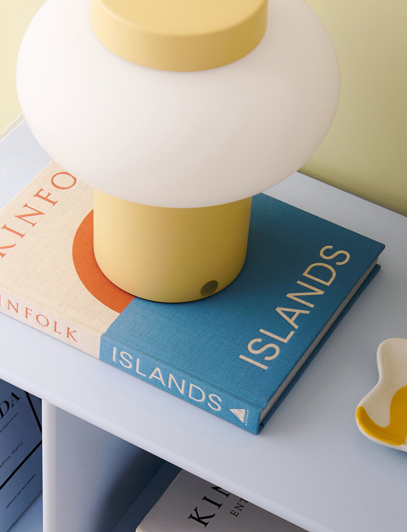 Gelb-weiße Pilzlampe auf zwei Büchern („KINFOLK“, „ISLANDS“) auf einem hellblauen Regal vor gelbem Wandhintergrund.