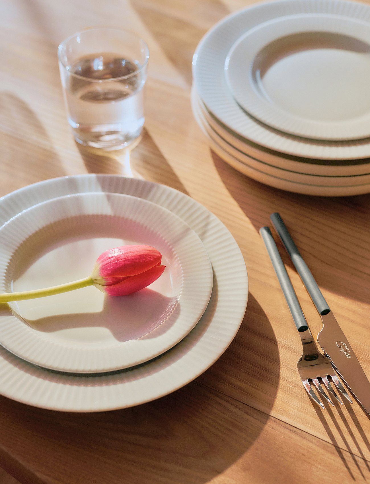Holztisch mit einem gedeckten Platz: weiße Teller, eine rosa Tulpe, ein Wasserglas und Besteck. Das Sonnenlicht wirft kräftige Schatten.