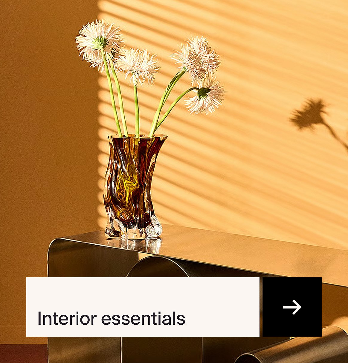 Swirled amber vase with light, fluffy flowers on a metallic table. Orange wall with blind shadows. "Interior essentials"