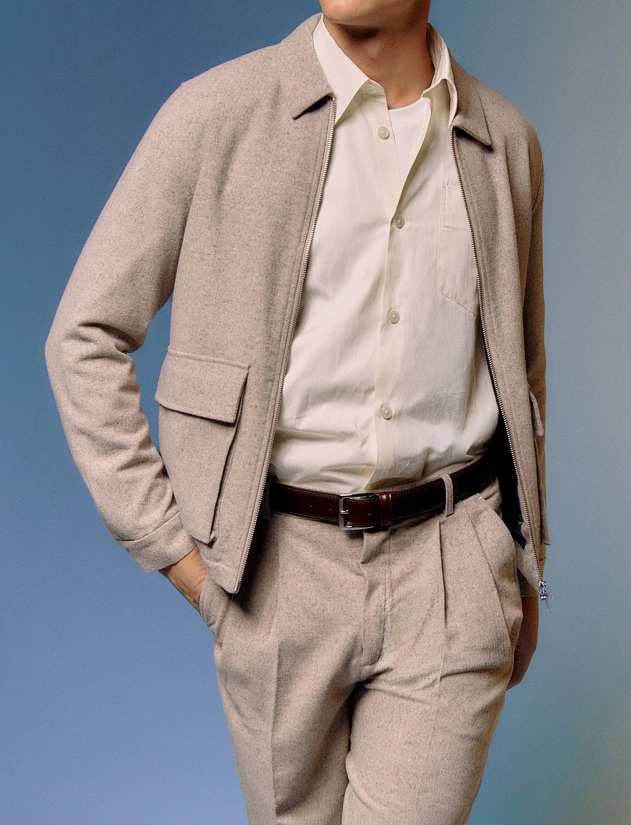 Person in a light beige textured jacket, cream shirt, brown belt, and matching pleated trousers. Blue background.