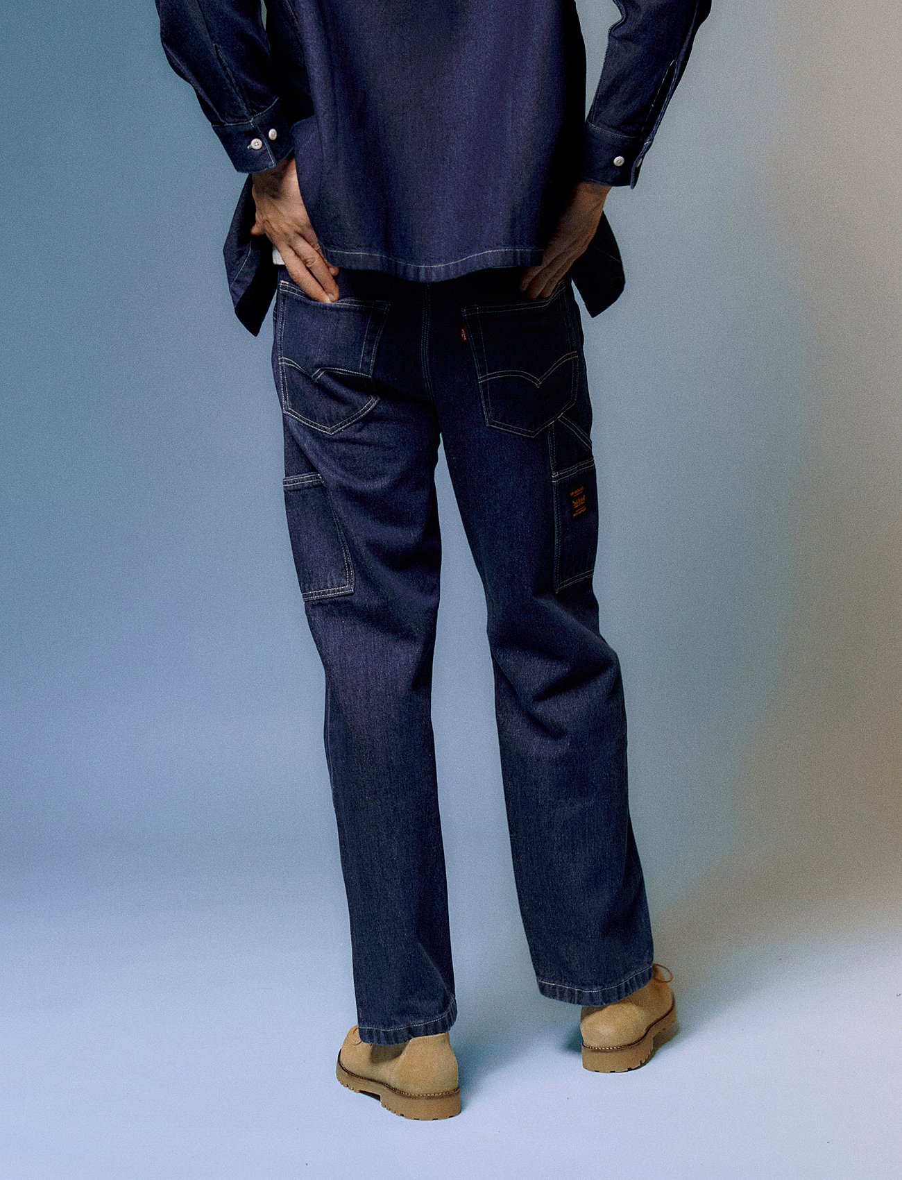 Person from behind in dark blue denim shirt and carpenter pants with tan shoes, hands in back pockets, light blue background.
