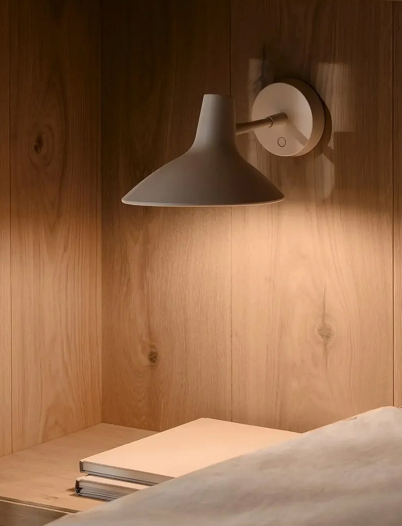Vaaleanharmaa seinävalaisin valaisee puuseinää ja hyllyllä olevia kahta kirjaa; etualalla on viltti.