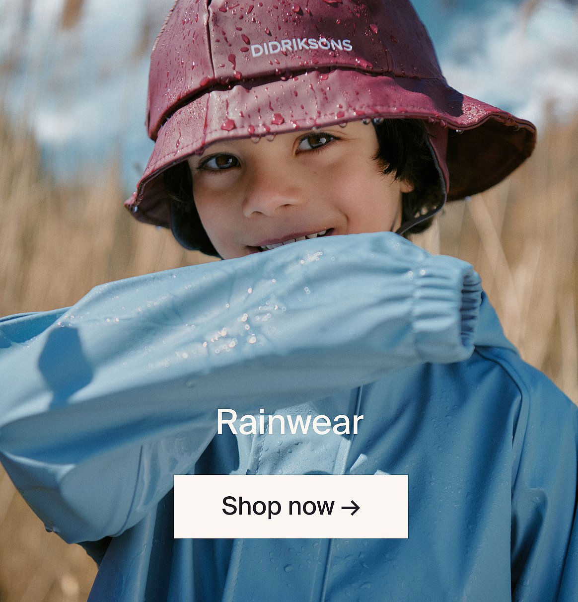 Smiling child in a maroon rain hat and blue rain jacket, covered in water droplets, outdoors. Text: Rainwear, Shop now.
