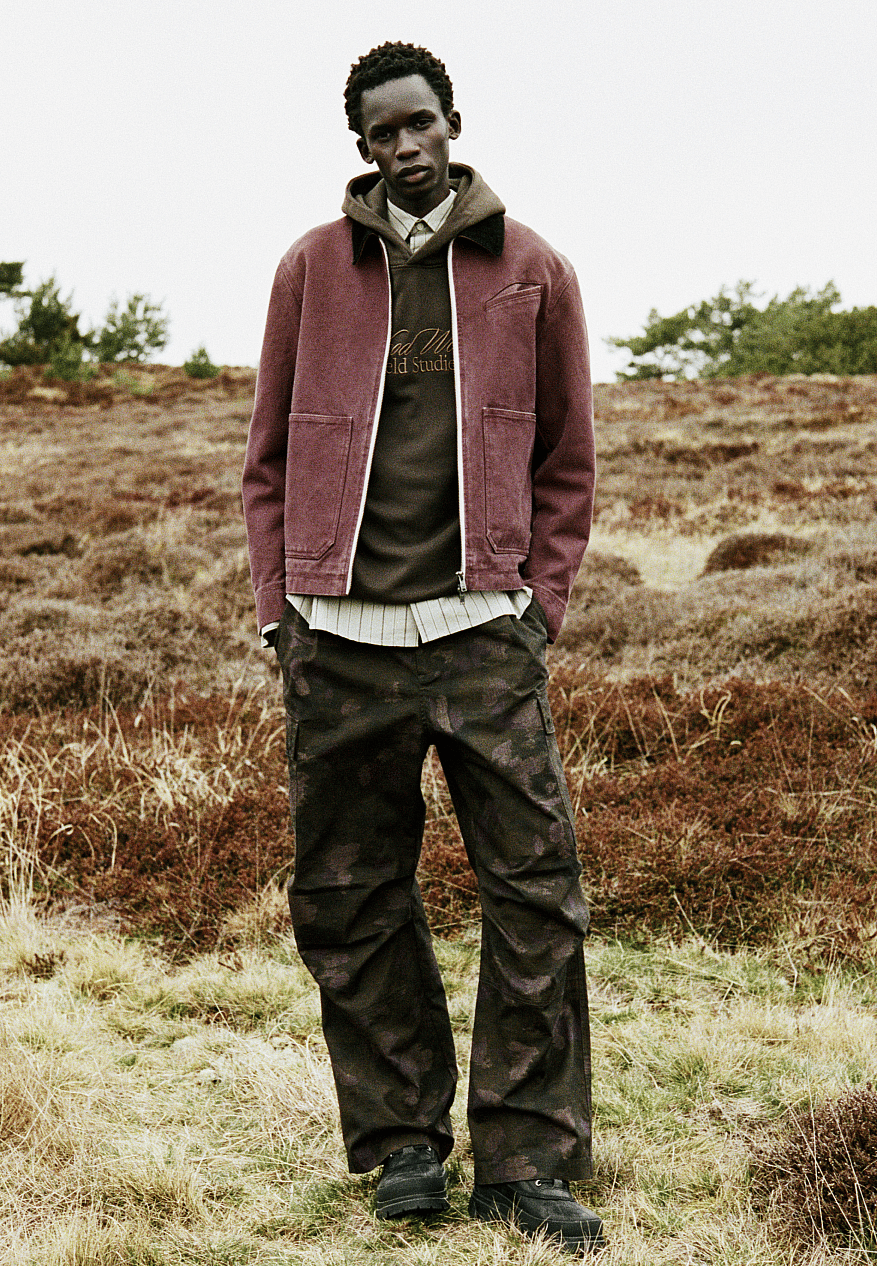 Black man in maroon jacket, olive hoodie, and dark camo pants stands in a sparse, brown and green field under an overcast