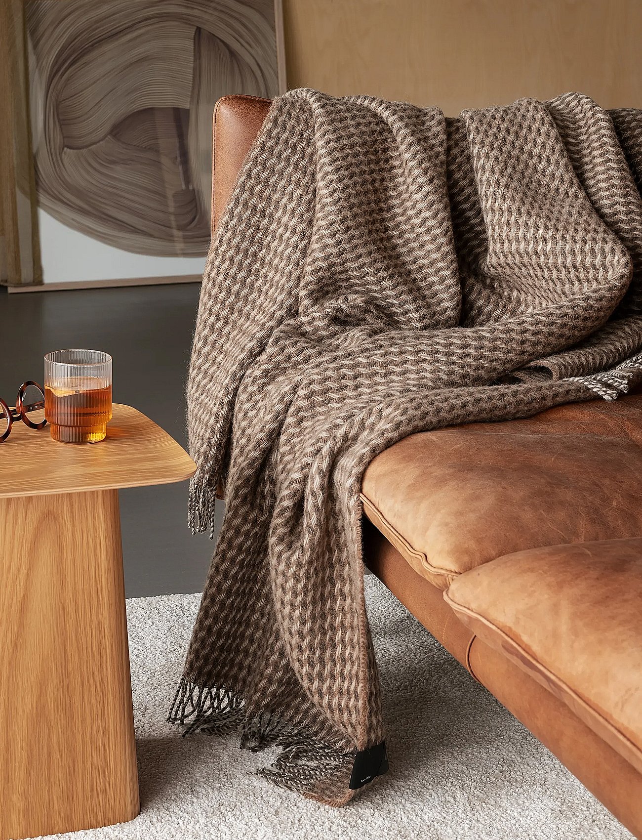Un plaid à motifs marron est posé sur un canapé en cuir. Une table d'appoint en bois accueille un verre et une paire de lunettes.