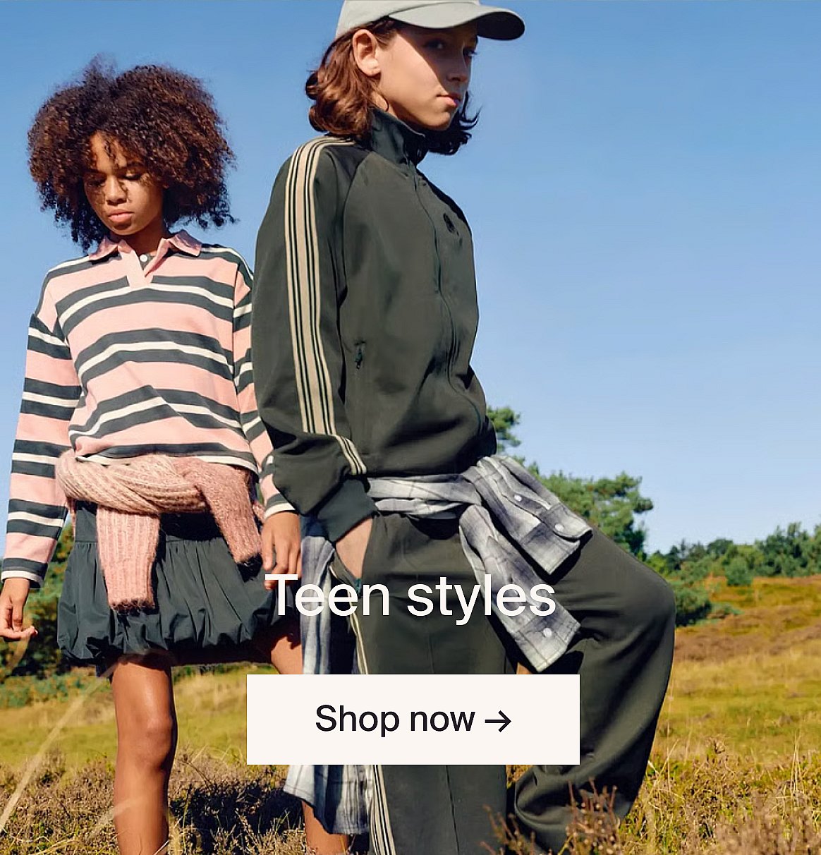 Two boys, one with curly hair in a striped polo, one in a red and blue jacket, sitting outdoors. Text: Teen styles, Shop now.