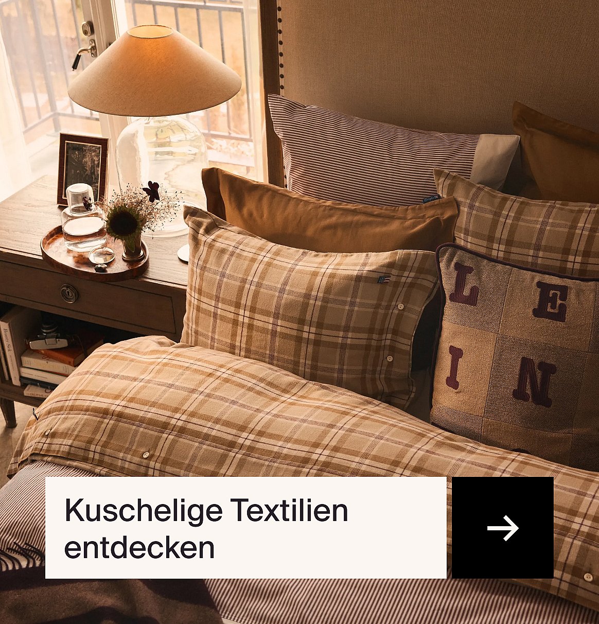 Ein gemütliches Bett mit karierter und gestreifter Bettwäsche, Kissen und einem Nachttisch mit einer Lampe, neben einem Fenster.