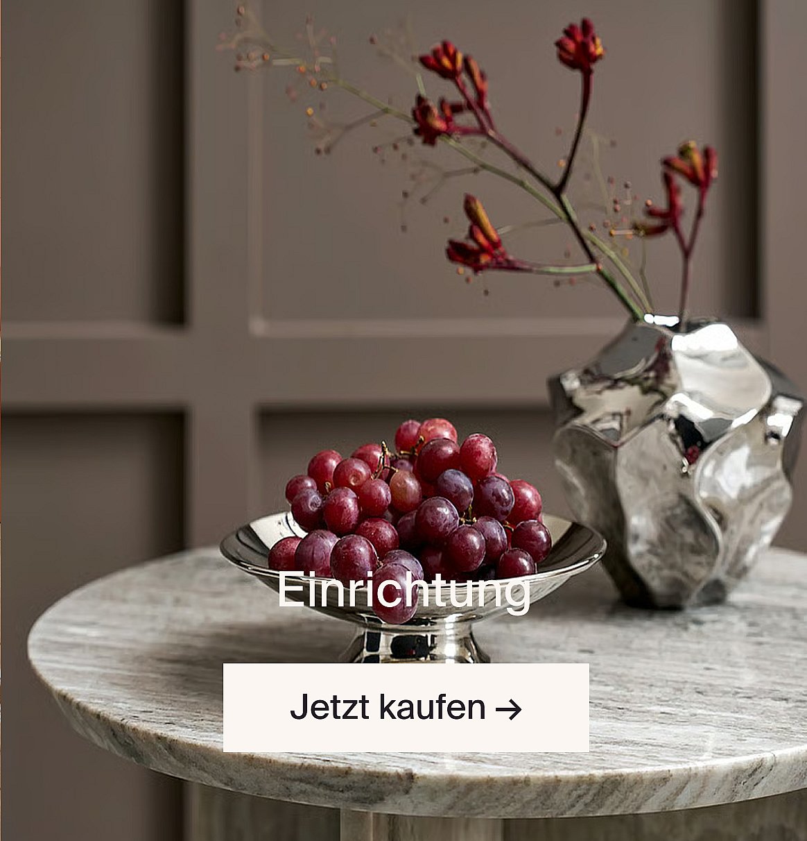 Rote Trauben in einer silbernen Schale und rote Blumen in einer silbernen Vase auf einem Marmortisch vor einer Wand mit braunen Vertäfelungen.
