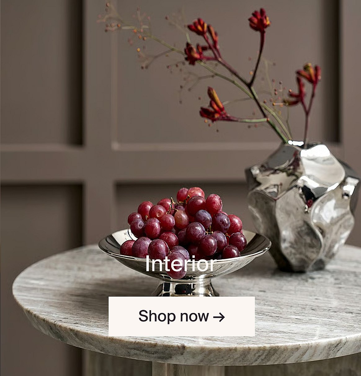 Red grapes in a silver bowl and red flowers in a silver vase on a marble table against a brown paneled wall.