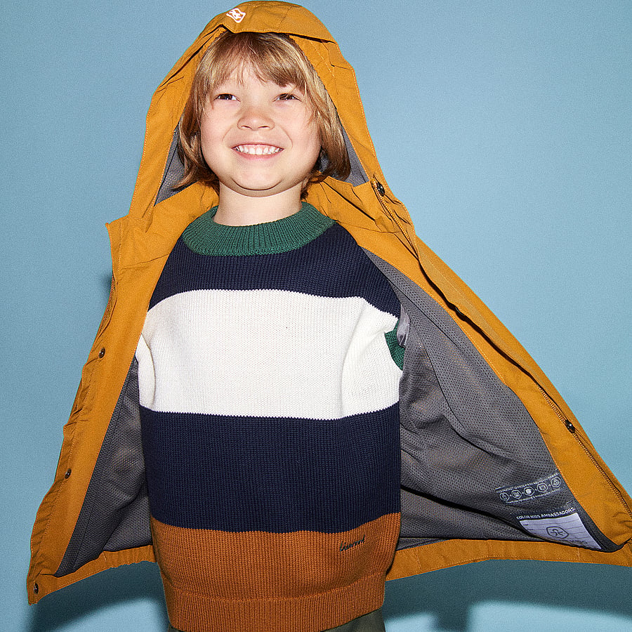 Child with an orange tiger backpack crouches on a white floor in front of a light blue wall. Backpacks.