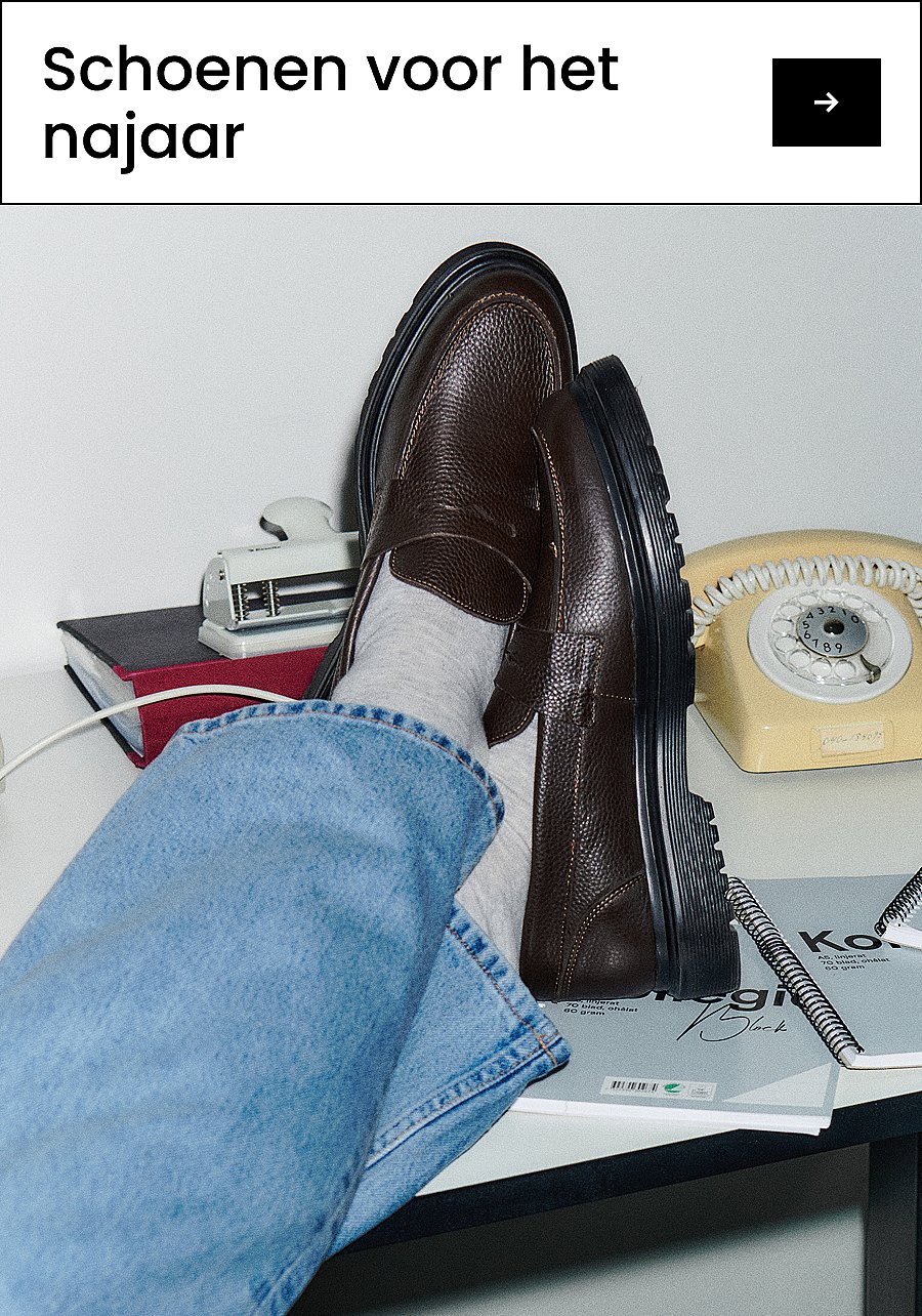 Benen in een donkere broek en lichte sokken, met bruine leren loafers met dikke zwarte zolen, staand op beton. Tekst: Schoenen
