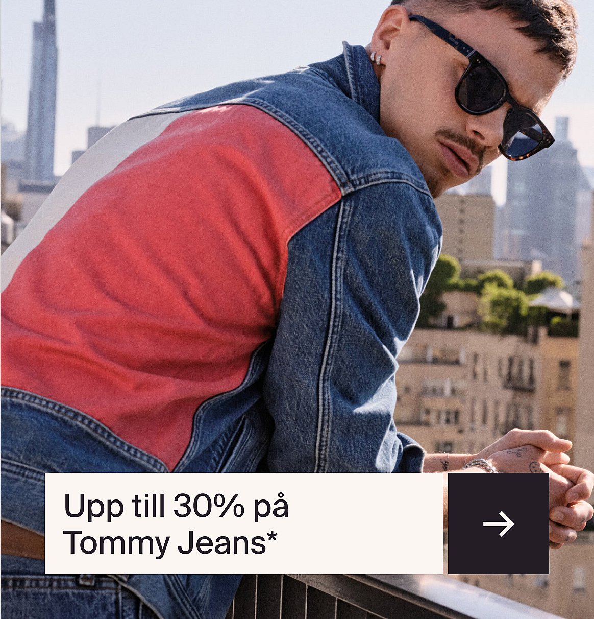 En man i en jeansjacka med färgblock och solglasögon, lutad mot ett räcke med stadens byggnader i bakgrunden.
