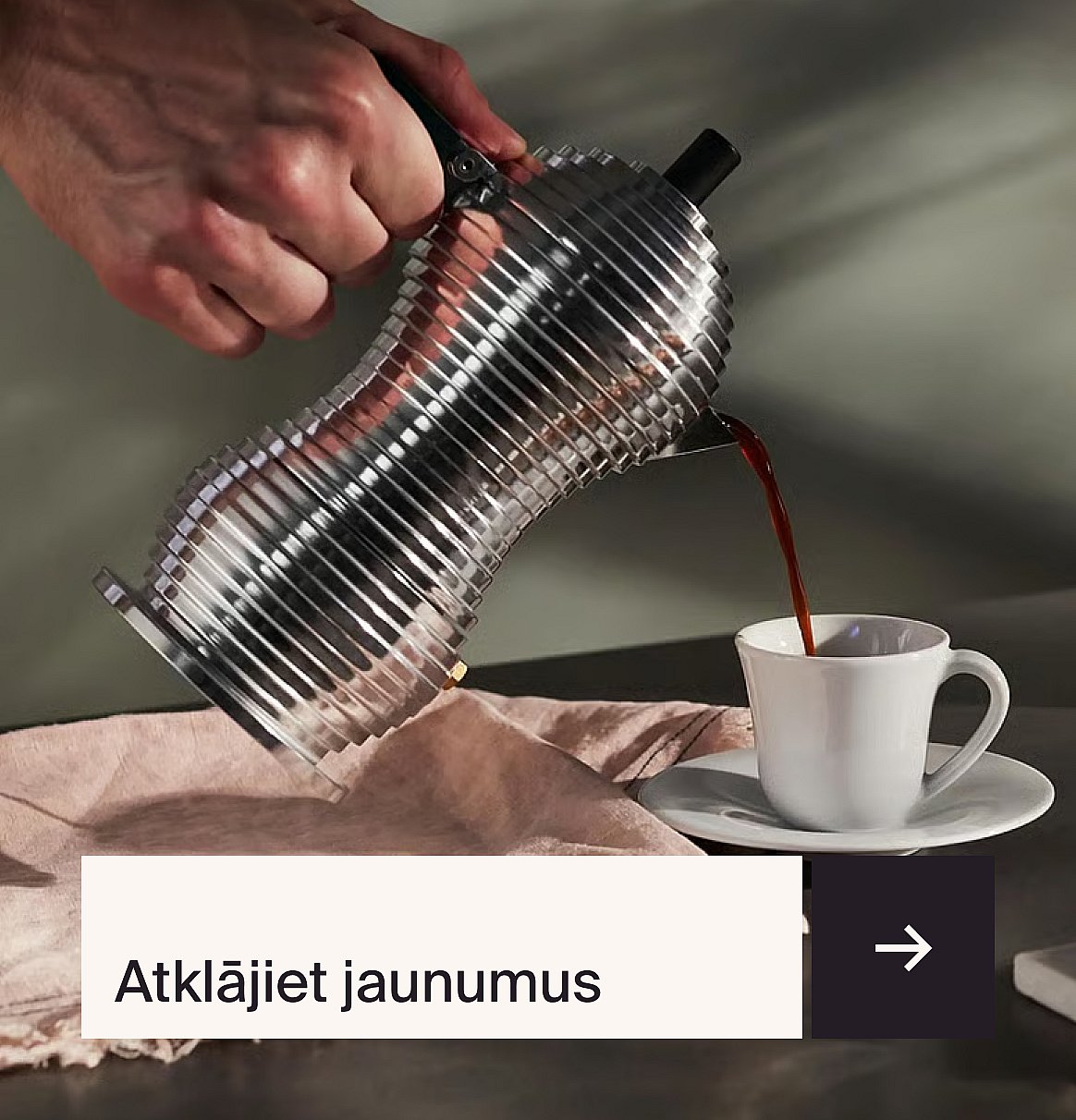 Kafija tiek ielieta ar rokām no rievotas metāla kafijas kannas baltā krūzē, kas stāv uz tumšas virsmas ar rozā audumu.