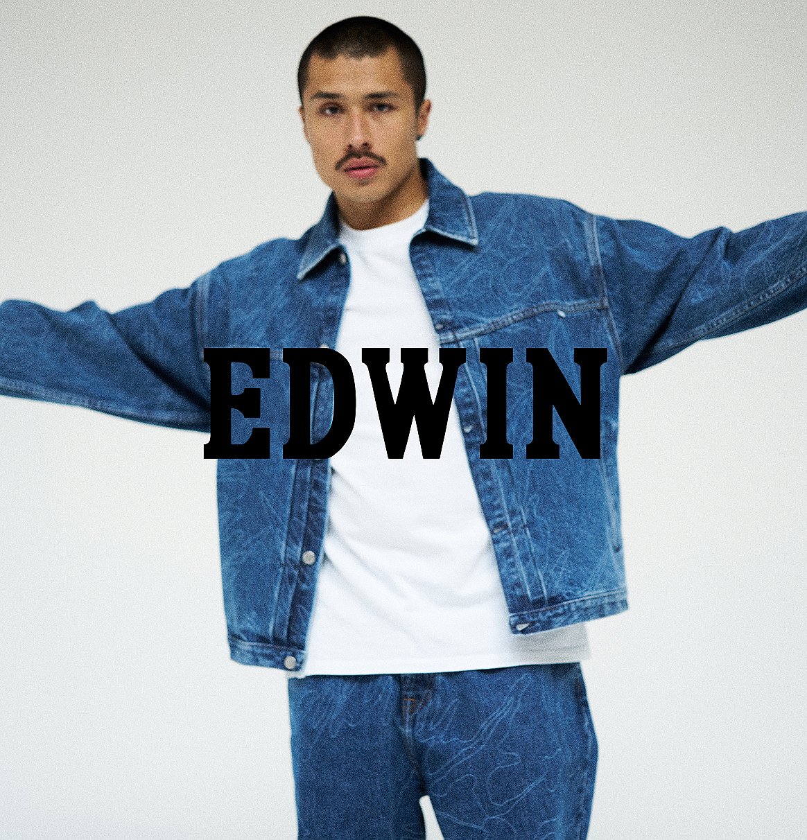 Man in a denim jacket and jeans, white t-shirt, mustache, arms outstretched on a white background. Text: EDWIN.