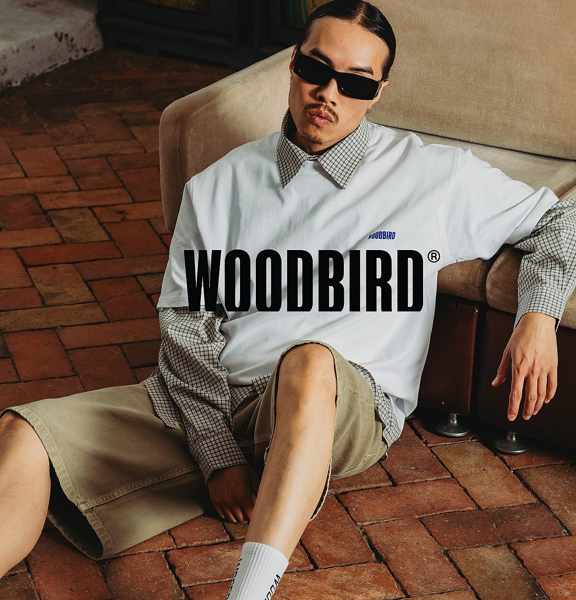 Man in sunglasses, white WOODBIRD tee over plaid shirt, khaki shorts, white socks, sitting on brick floor by sofa.