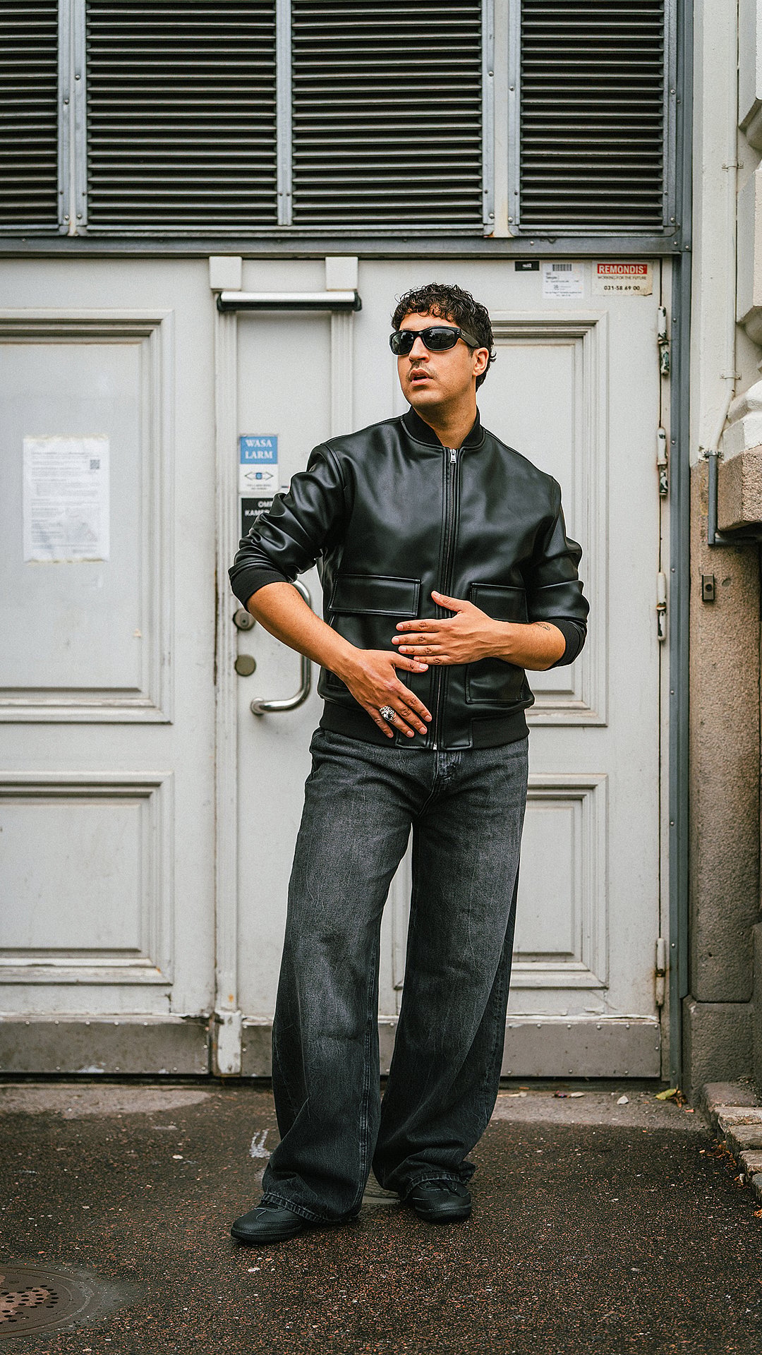 Man in black leather bomber jacket, dark wide-leg jeans, and sunglasses stands looking left in front of a gray door.